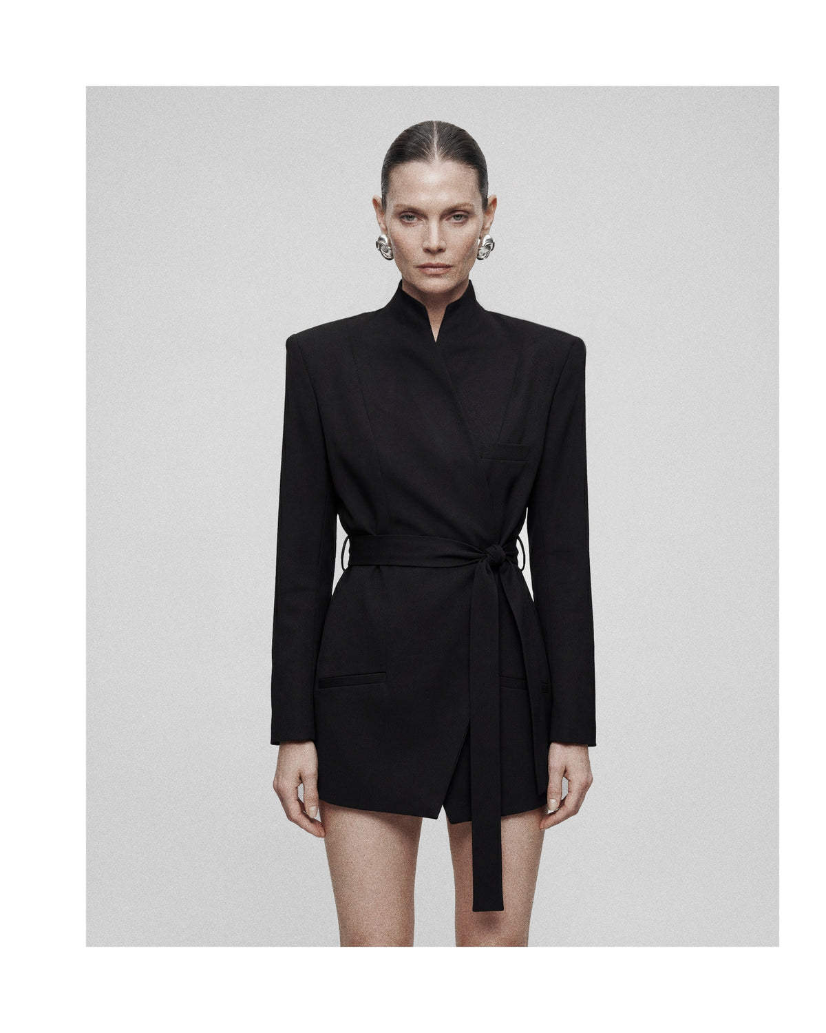 A model wearing a black long-sleeve wrap-style mini dress with a high stand collar, structured shoulders, and a self-tie belt, set against a solid light gray background.