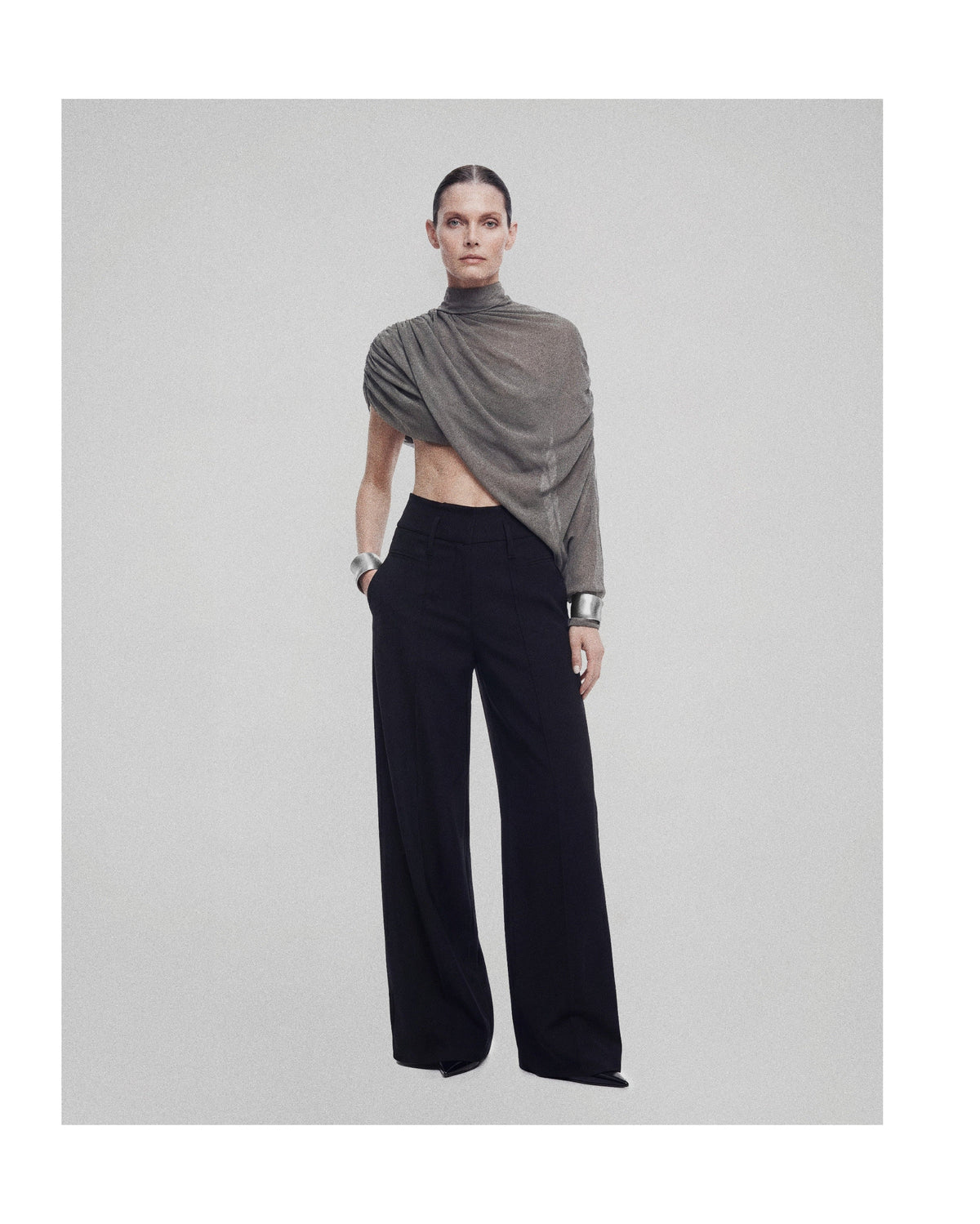 A model stands against a light gray studio background wearing a draped taupe-gray asymmetrical top and black wide-leg trousers.