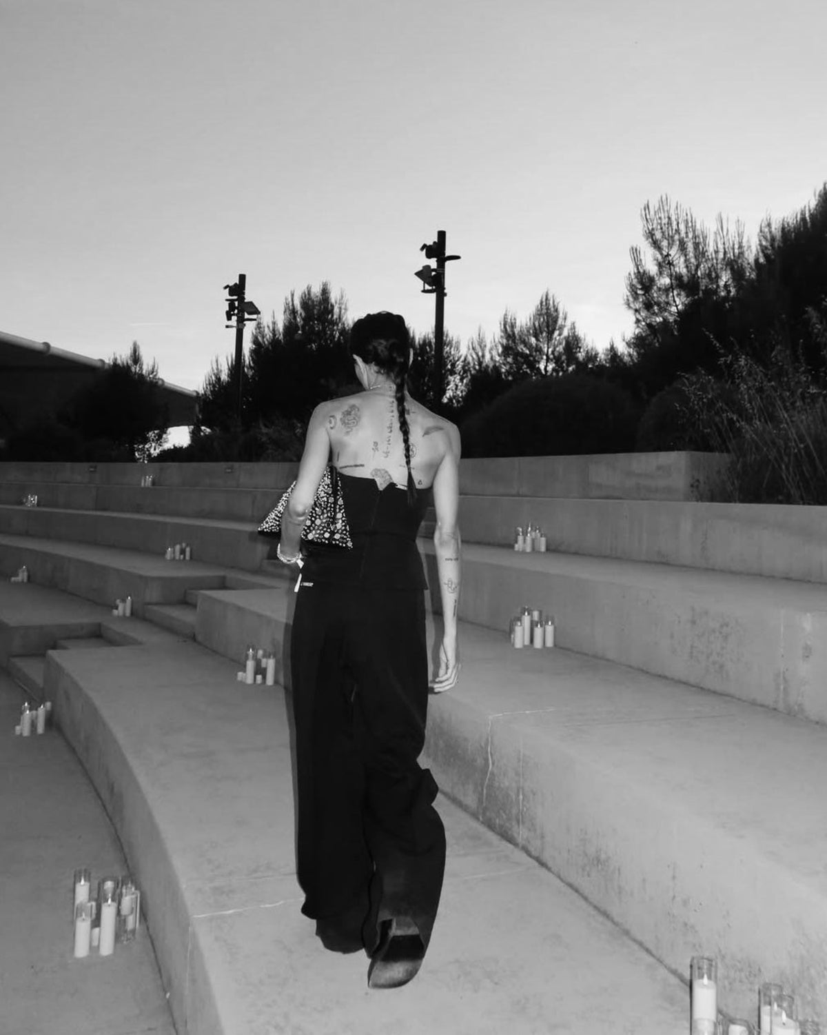 A black and white photograph of a model from behind walking down concrete outdoor steps, wearing a dark strapless jumpsuit and carrying a patterned handbag.