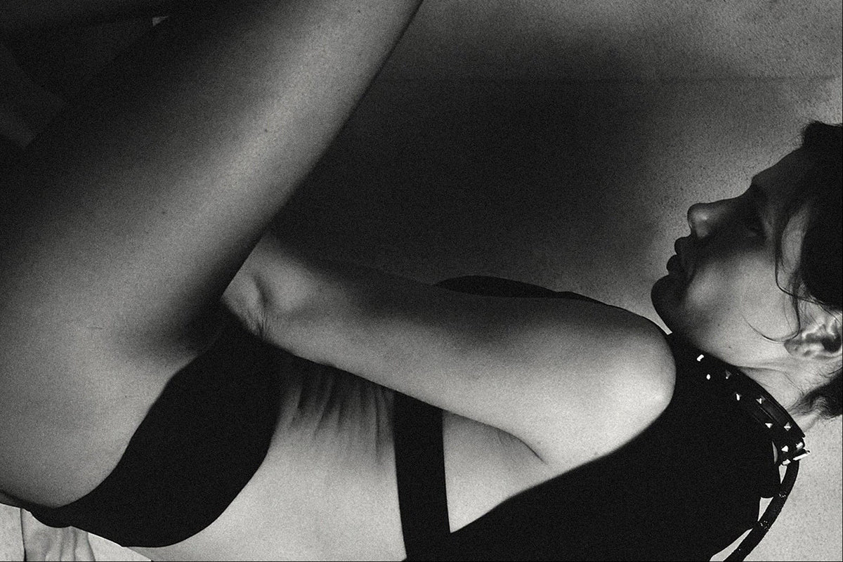 A black and white four-panel fashion photograph showing a seated nude model from different angles, wearing a black strappy garment and sheer black tights against a textured studio backdrop.